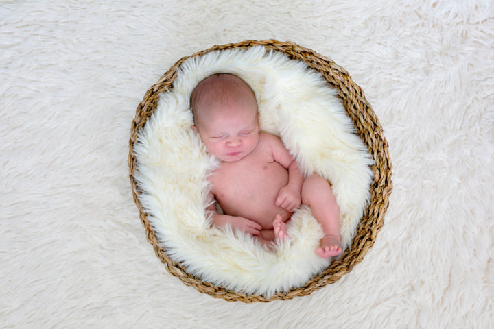 A naked new baby cuddled up on a fluffy white blanket in a wicker basket